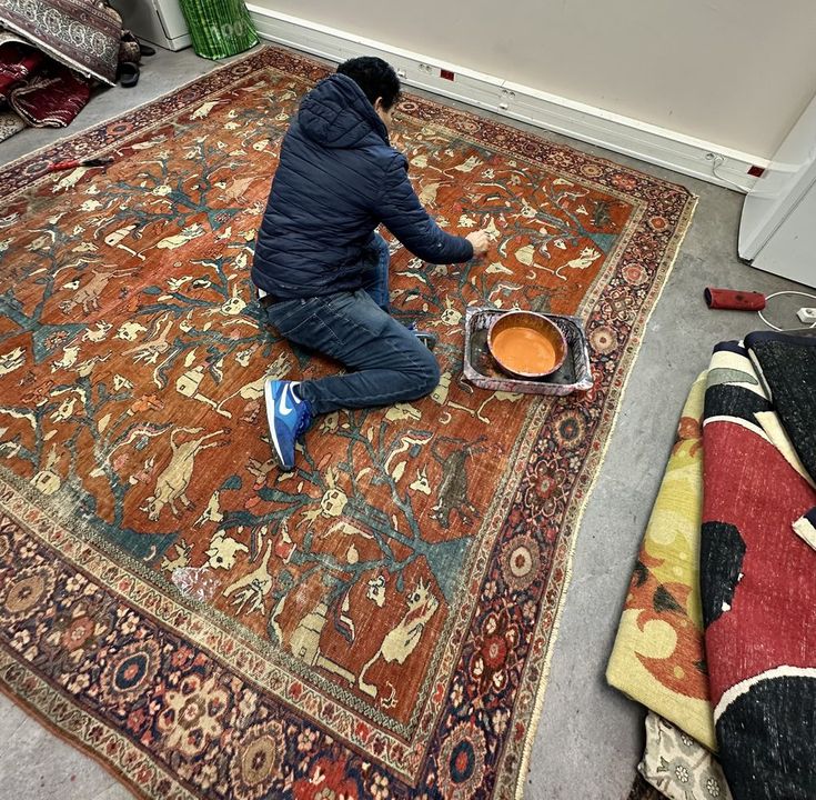 A craftsman kneeling on a large Persian rug with an animal design, re-dyeing the red field with a brush