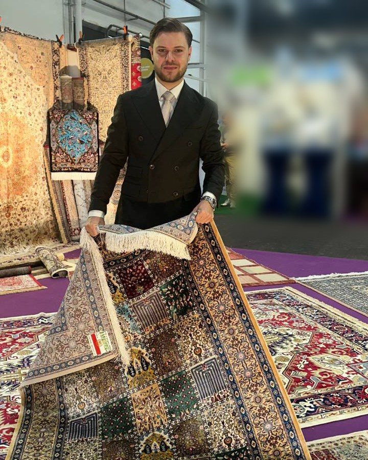 Adrien Mignaton, en costume, présente un tapis persan en soie sur un salon, devant d'autres tapis d'Orient
