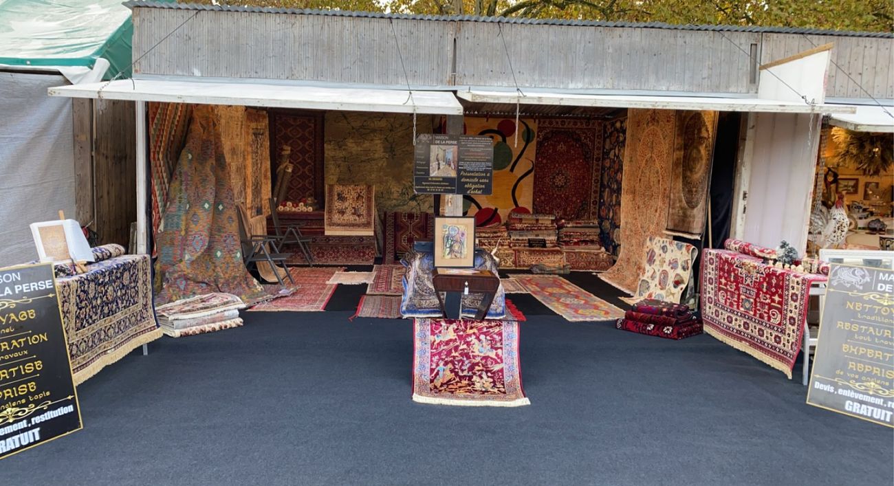 La Maison de la Perse's open-air stand: Oriental rugs hanging and laid on the ground, with signs reading “Cleaning, restoration, valuation, trade-in”