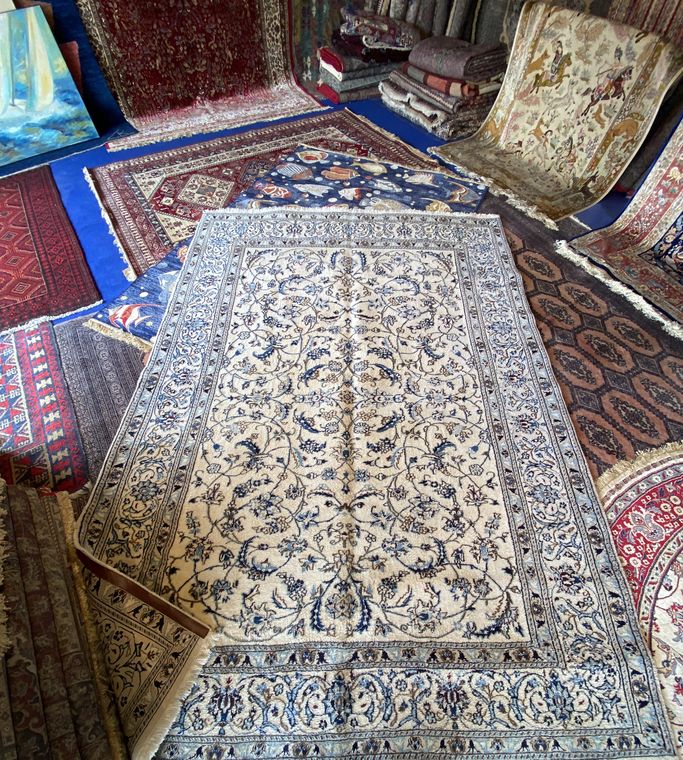 Persian Naïn rug in wool and silk, with an ivory ground and blue floral scrolls, unfolded among other rugs from our stock